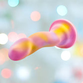 The Avant Sun's Out Dildo by Avant is a pastel pink silicone dildo with abstract curves and yellow and purple swirls, featuring a wide, rounded base for harness compatibility, shown against a softly lit, blurred background.